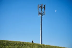 برج سلولی روی یک تپه با یک نفر ایستاده در زیر و یک ماه روز در آسمان.