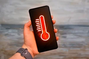 دستی که یک گوشی هوشمند را با دماسنج در دست گرفته و نشان می دهد که دستگاه در حال داغ شدن بیش از حد است.